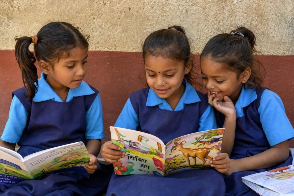 Three young village schoolgirls sitting against a wall reading colorful picture books together natural daylight | edCircuit Three young village schoolgirls sitting against a wall reading colorful picture books together natural daylight