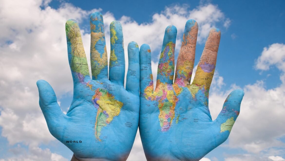 Two open palms painted with a colorful world map of the continents held up against a bright blue sky with white clouds