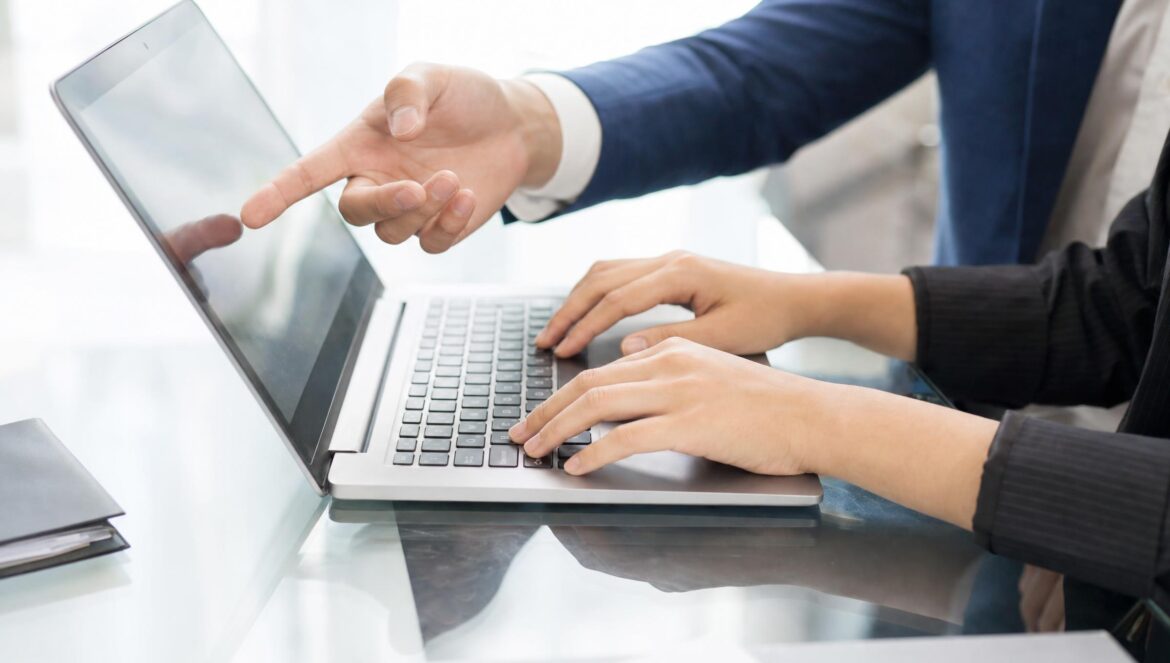 Two people at a glass desk one typing on a laptop while the other points at the screen closeup on hands bright modern office light | edCircuit Two people at a glass desk one typing on a laptop while the other points at the screen close up on hands bright modern office light