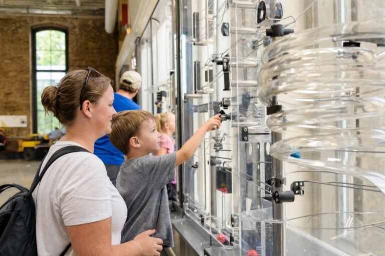 Visitors at a hands-on science museum exhibit, touching a tall clear panel of rolling ball tracks and coiled tubes in a bright gallery