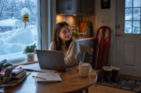 Snow days a girl does schoolwork on a laptop at the kitchen table as snow buries the school bus stop sign outside