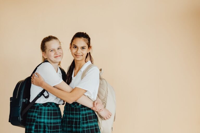 Two schoolgirls wearing uniforms and backpacks, hugging and smiling in a bright indoor setting