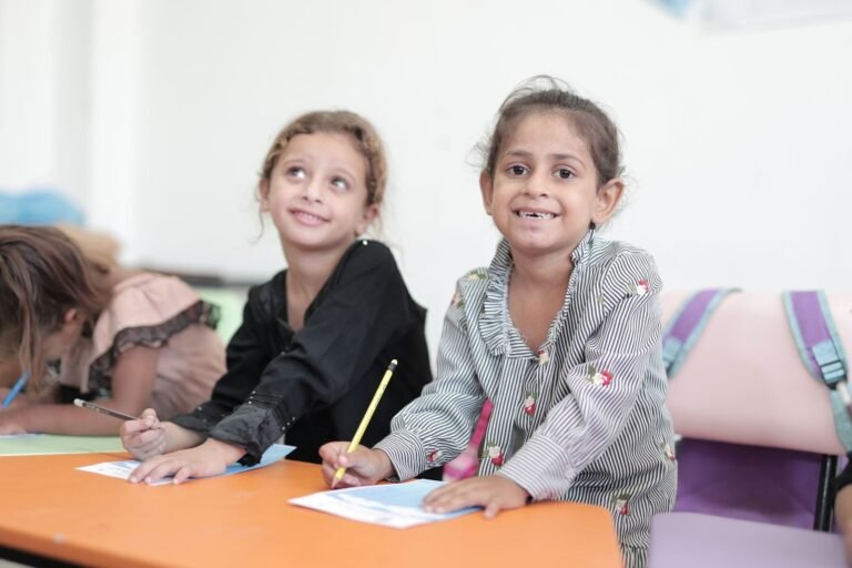 Two young children smiling while engaging in a classroom activity with pencils and paper