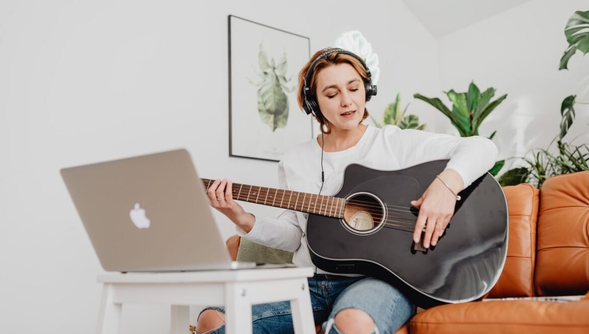 Woman playing guitar with headphones in a cozy living room using lapt