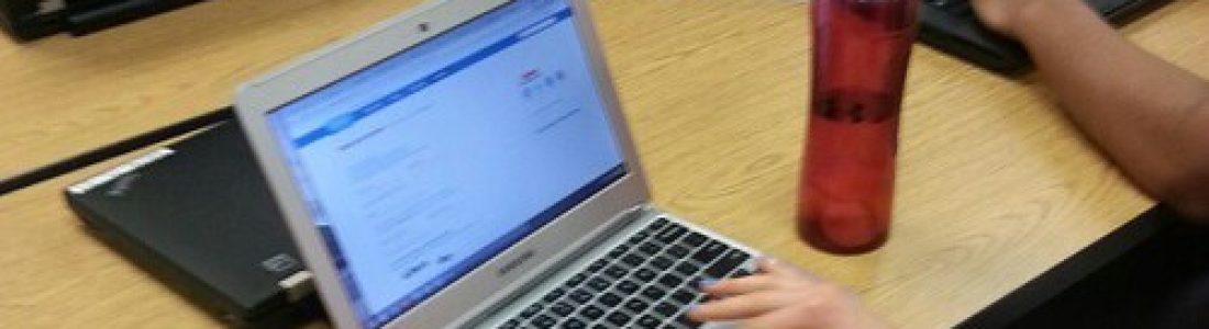 Students sit at a table with laptops, working on assignments. One student in the foreground types on a silver laptop next to a red water bottle. Papers and more laptops are scattered on the table.