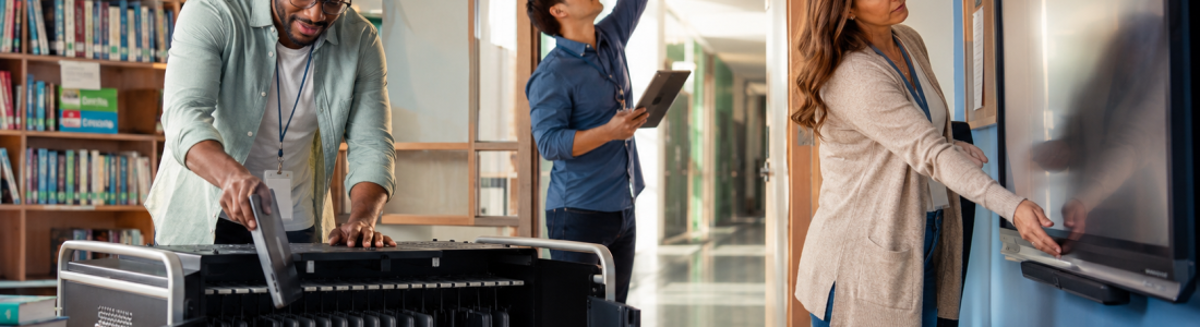 Three adults in a library: man loading a large printer, another man holding a tablet and reaching the ceiling sensor, woman using a touchscreen board.