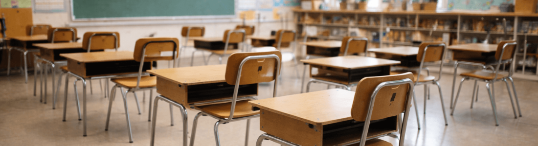 Rows of empty wooden desks and chairs in a well-lit classroom with a green chalkboard and educational posters on the walls reflect the impact as School District Layoffs Surge Nationwide.