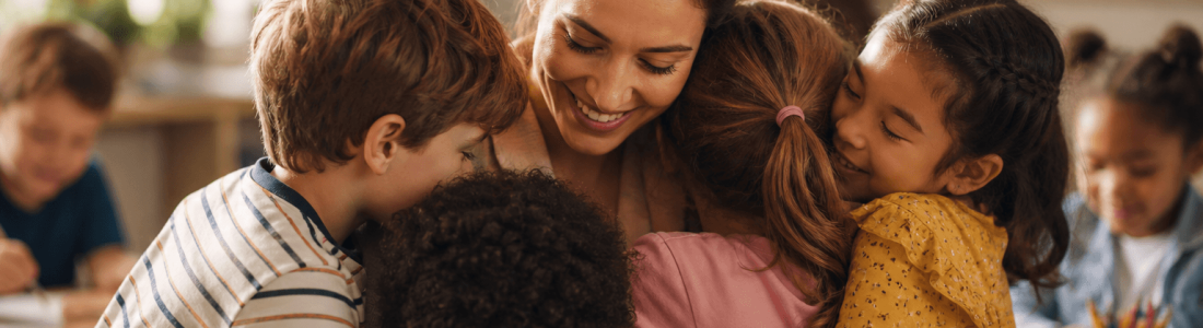 A teacher sits in a classroom as four young students hug her, surrounded by other children and colorful classroom decorations, capturing the warmth of Teacher Appreciation Week.