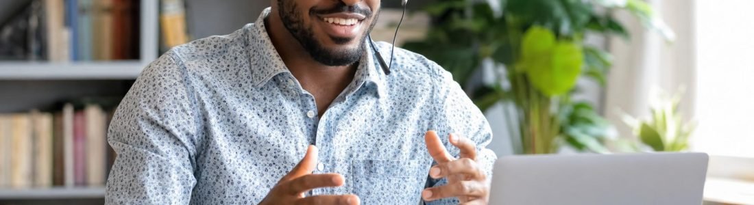 A smiling Black man wearing a headset and glasses gesturing as he talks on a video call at a laptop in a bright home office with plants