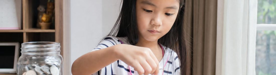 A young girl stacking coins into neat piles on a table next to a glass jar of coins, soft daylight