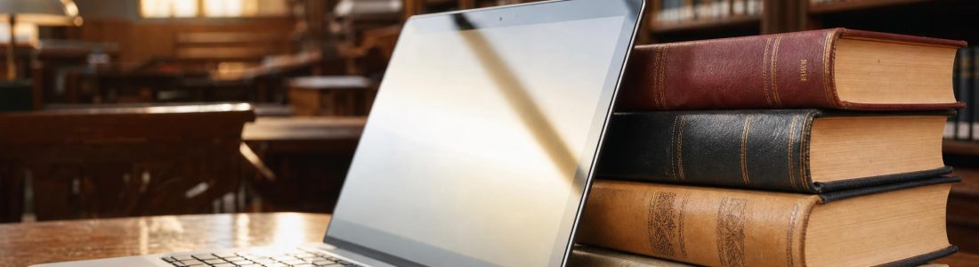 An open laptop leaning against a stack of hardcover books in a library, warm golden window light