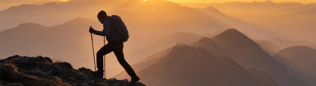 The silhouette of a hiker with a backpack and walking poles climbing a mountain ridge at sunrise, layered hazy mountains behind