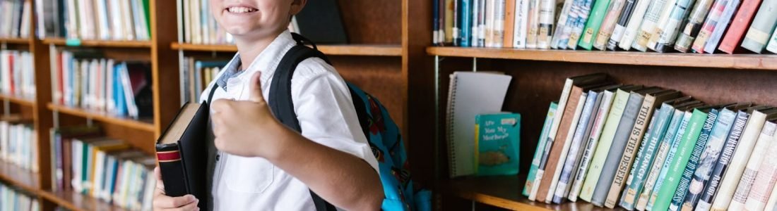 Young boy in a library giving a thumbs up while holding a book
