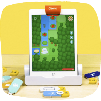 A tablet on a stand displays a colorful, cartoon-like game with trees, water, and a character. The tablet has an orange Osmo clip on top. Various game pieces are scattered on the wooden surface in front.