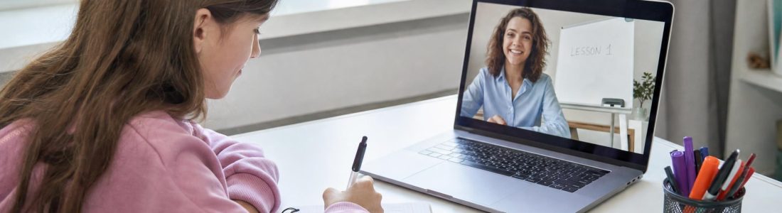 A girl writing in a notebook at a white desk while a smiling teacher appears on a video call on her open laptop, bright daylight from a window