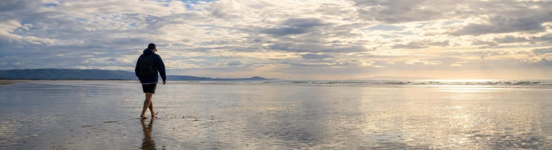A lone adult walking across a wide wet sandy beach at low tide that mirrors a bright cloudy sky, sun breaking through the clouds