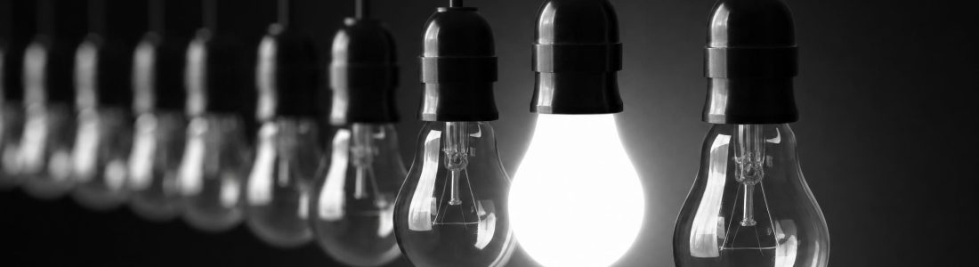 A row of clear light bulbs hanging on black cords against a dark background, all unlit except one glowing bright white bulb, moody black and white tones