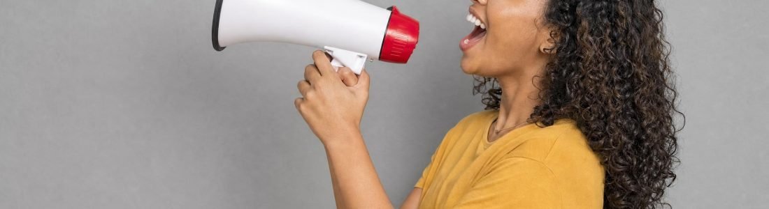 A smiling woman with curly hair shouting into a white megaphone against a plain gray wall, soft studio light