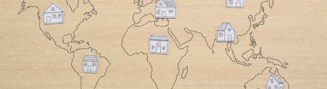 A world map outline drawn in thin ink on a light wooden tabletop with small house sketches placed across the continents, even overhead light