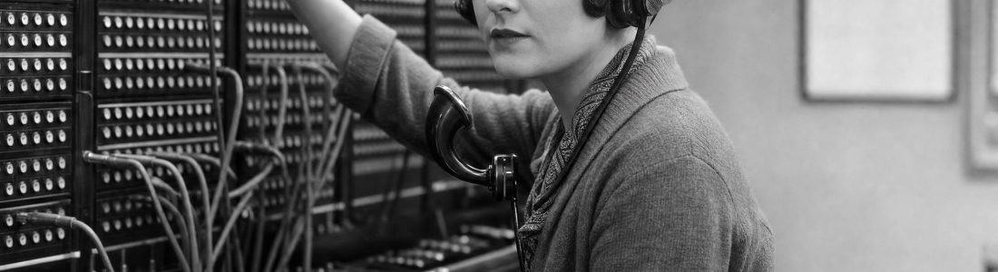 A young woman in a 1920s cardigan working at an old telephone switchboard, plugging patch cords into a wall of jacks while wearing a vintage headset, black and