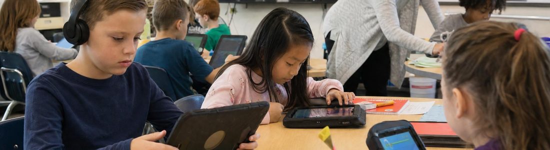 Elementary students at classroom tables playing a learning game on tablets, one wearing headphones, a teacher helping in the background and a wall screen showin