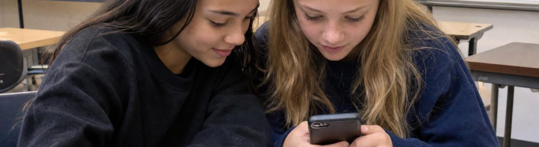 Two middle school girls sitting at a classroom desk looking down at a cell phone together, notebooks open in front of them, indoor light
