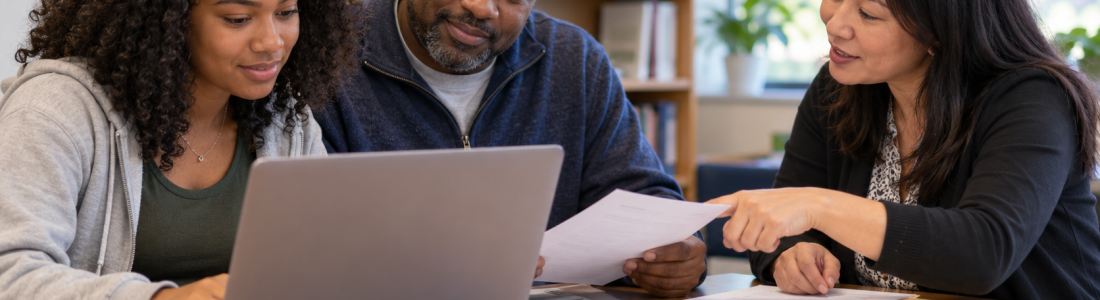 Three colleagues sit at a table, looking at a laptop and discussing printed documents in a library setting.