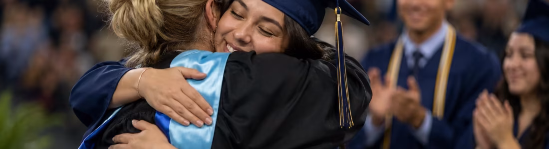 Graduate in a blue cap and gown hugs a smiling person at a graduation ceremony as classmates applaud in the background of a crowded arena.