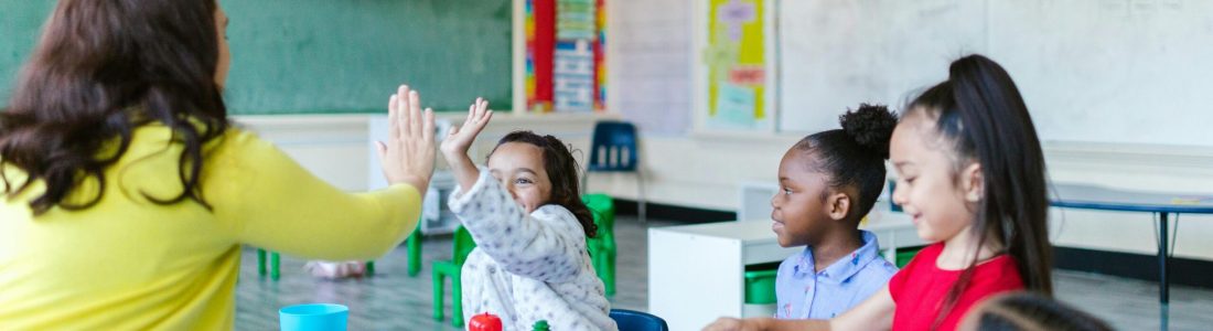 A teacher gives a high five to a smiling young girl at a table with three other children in a colorful classroom. The children are playing with educational toys and art supplies.