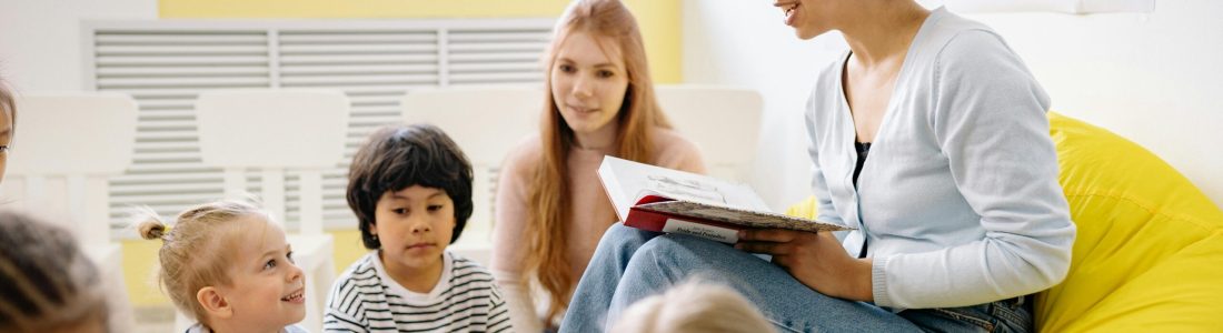 A woman sits on a yellow beanbag reading a book to a group of young children, with another adult nearby. The children sit and listen attentively in a bright classroom with alphabet posters on the wall.