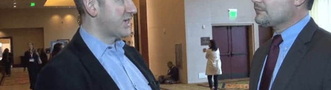 Two men in business attire are having a conversation in a spacious, well-lit hallway with patterned carpet and round hanging lights. Other people can be seen in the background near large doors.